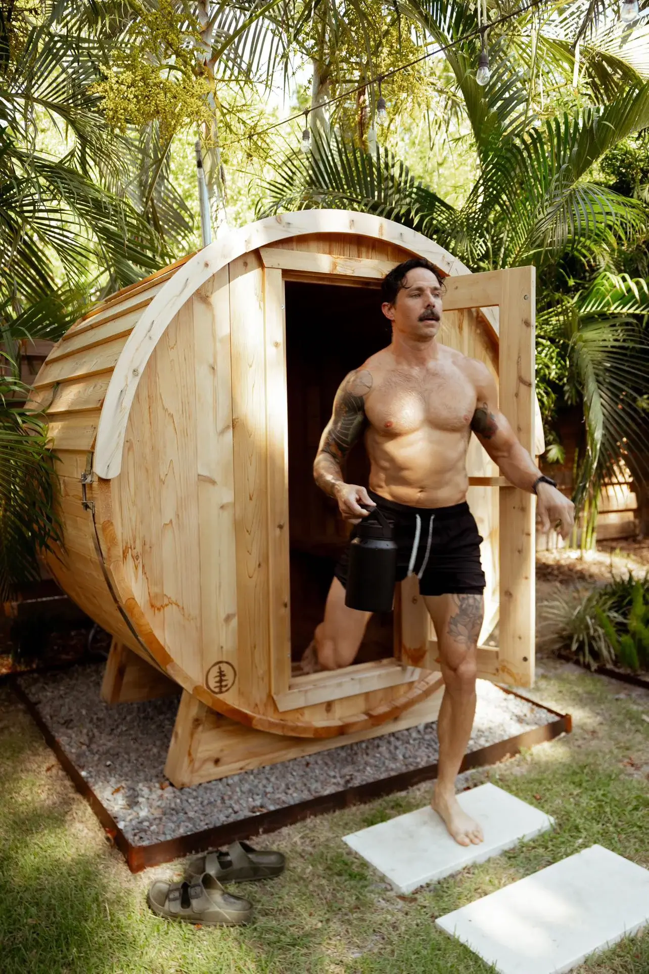 A man stepping out of a barrel sauna carrying a bucket
